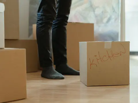 A cardboard box with Kitchen written on its side in red marker, on a wood floor among other stacked moving boxes, with a person's legs behind it