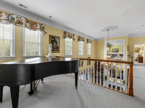 A black grand piano on a carpeted upstairs landing beside a wooden balustrade, with tall windows along one wall and an open doorway beyond