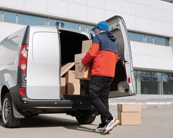 A person in an orange and navy jacket and a blue knit cap, seen from behind, lifts cardboard boxes into the open back of a silver van, with two more boxes on the pavement