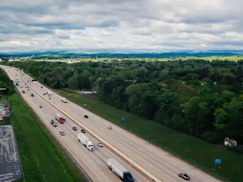 Tractor-trailers and cars on a six-lane divided interstate running through wooded green country under a cloudy sky, seen from above
