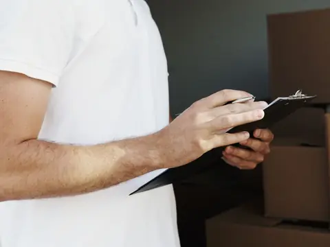 A man in a white T-shirt, shown from the shoulders down, writes on a black clipboard in front of stacked cardboard boxes