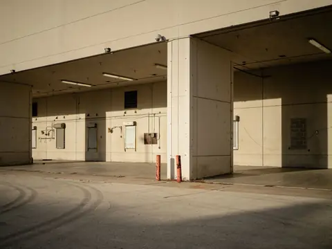 An empty covered loading dock with roll-up doors, dock lights and bollards along the back wall of a large concrete building