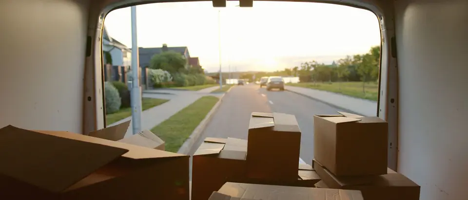 Cardboard boxes in the back of a van, looking out through its open rear doors onto a residential street lined with lawns and houses at sunset