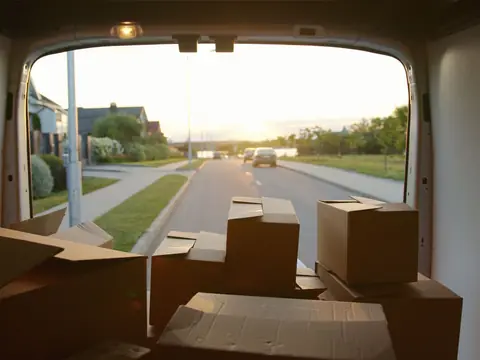 Cardboard boxes in the back of a van, looking out through its open rear doors onto a residential street lined with lawns and houses at sunset