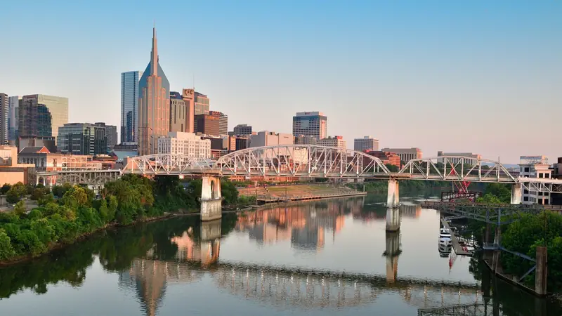 Downtown Nashville at sunrise, with the twin-spired tower known as the Batman Building above the John Seigenthaler Pedestrian Bridge and the skyline mirrored in the Cumberland River
