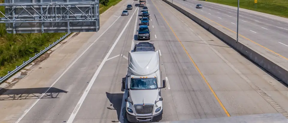 A white tractor-trailer leads a line of cars on a divided interstate between wooded green hills, seen from an overpass under a blue sky