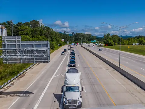 A white tractor-trailer leads a line of cars on a divided interstate between wooded green hills, seen from an overpass under a blue sky
