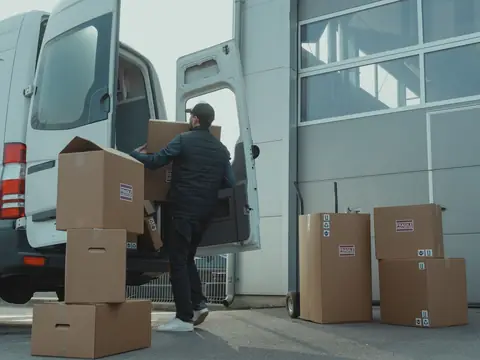 A man in a dark quilted vest and cap, seen from behind, lifts a cardboard box into the back of a white van, with more boxes stacked on the pavement beside a hand truck