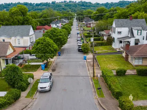 A straight residential street lined with trees and older frame houses, with cars parked along one curb and a wooded ridge behind, seen from above