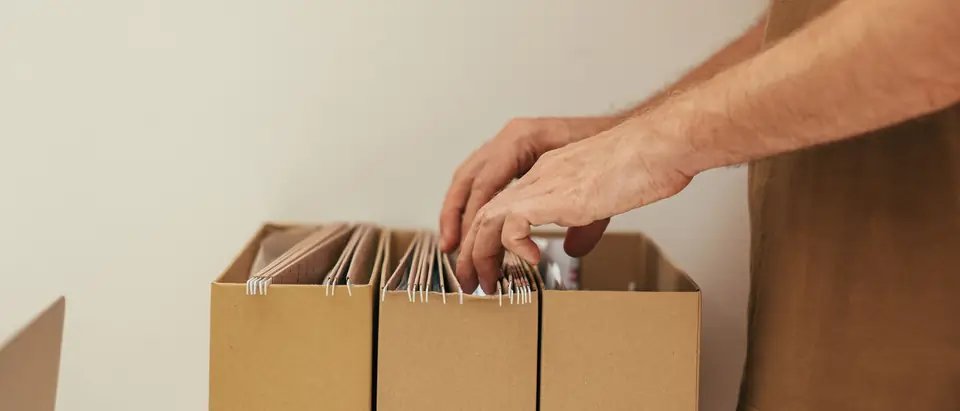 Two hands leafing through hanging file folders packed in three cardboard file boxes