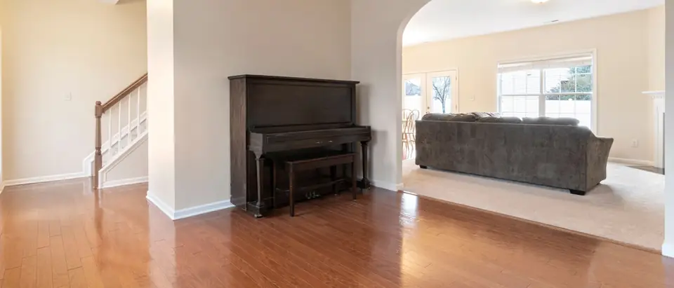 A dark upright piano and its bench against a wall in an empty room with hardwood floors, between a staircase and an archway to a carpeted living room