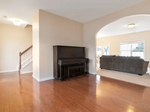A dark upright piano and its bench against a wall in an empty room with hardwood floors, between a staircase and an archway to a carpeted living room