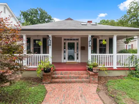 Brick steps and a brick walk leading up to the covered front porch of an older one-story house with white railings and a red porch floor