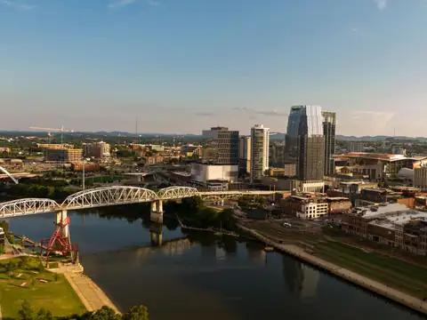 Downtown Nashville seen from above the east bank of the Cumberland River, with the white truss pedestrian bridge crossing to the riverfront and towers behind