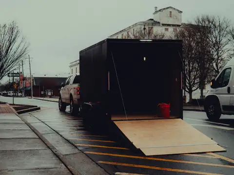 An enclosed cargo trailer hitched to a pickup at the curb of a wet small-town street, with its rear ramp lowered to the pavement
